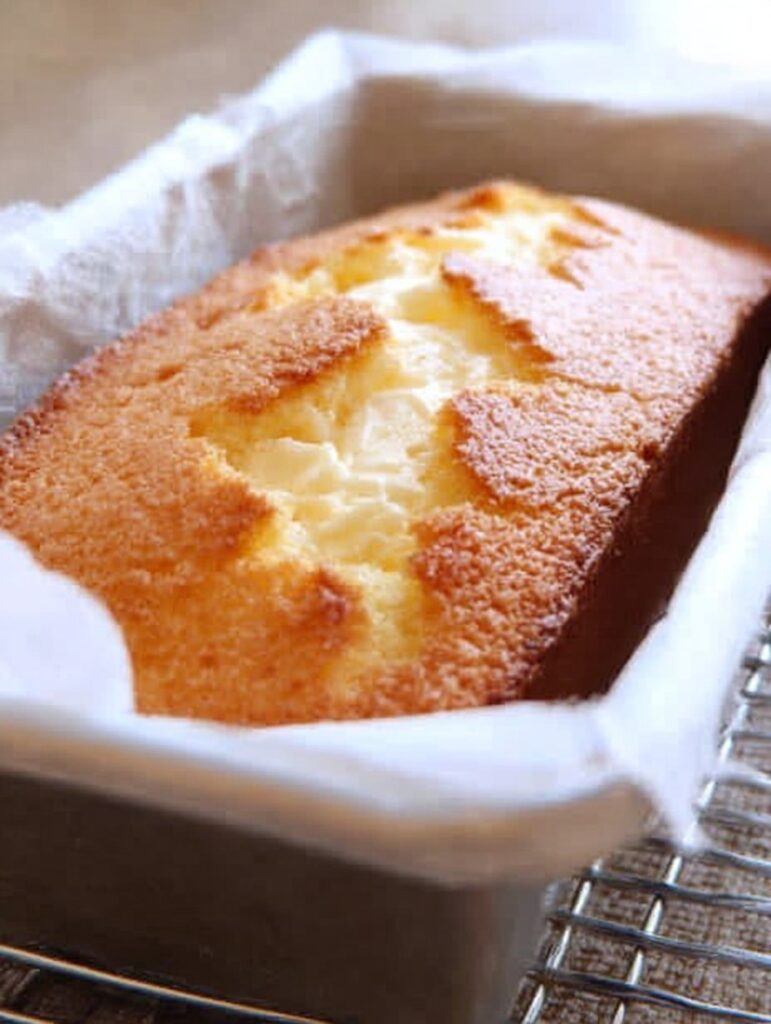 Panqué de queso crema dorado, servido en rebanadas sobre una tabla de madera, con un fondo acogedor y una taza de café al lado.